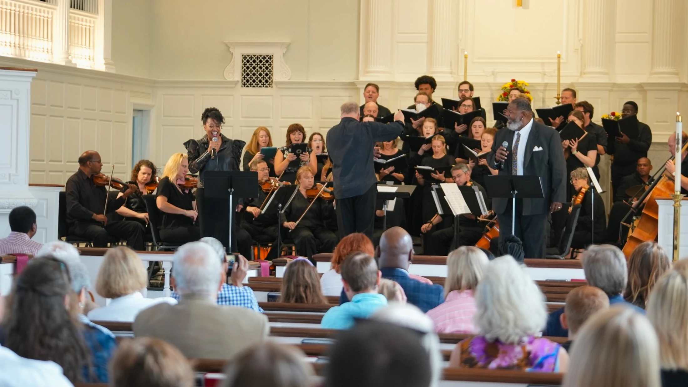A choir, orchestra, and two vocal soloists perform on stage in a bright hall for an audience seated in pews. The conductor is directing the musicians.